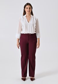 Donna in una blusa bianca trasparente con volant, pantaloni borgogna su misura e scarpe burgogna a punta, davanti a uno sfondo grigio neutro.