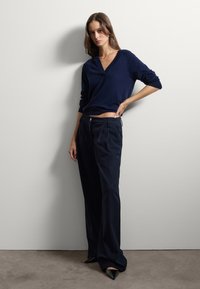 Pull en V bleu marine à manches longues, associé à un pantalon taille haute sombre et des talons aiguilles noirs. Tissu lisse, détails minimalistes.