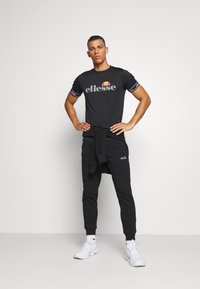 Black t-shirt with logo, striped sleeves; black sweatpants with cuffed hems, logo accent; white athletic shoes. Clean, sporty aesthetic.