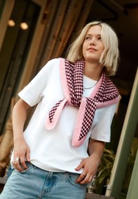 Écharpe tricotée à motif pied-de-poule rose et marron, portée sur un t-shirt blanc, avec deux longues lanières. Un jean en denim complète la tenue.