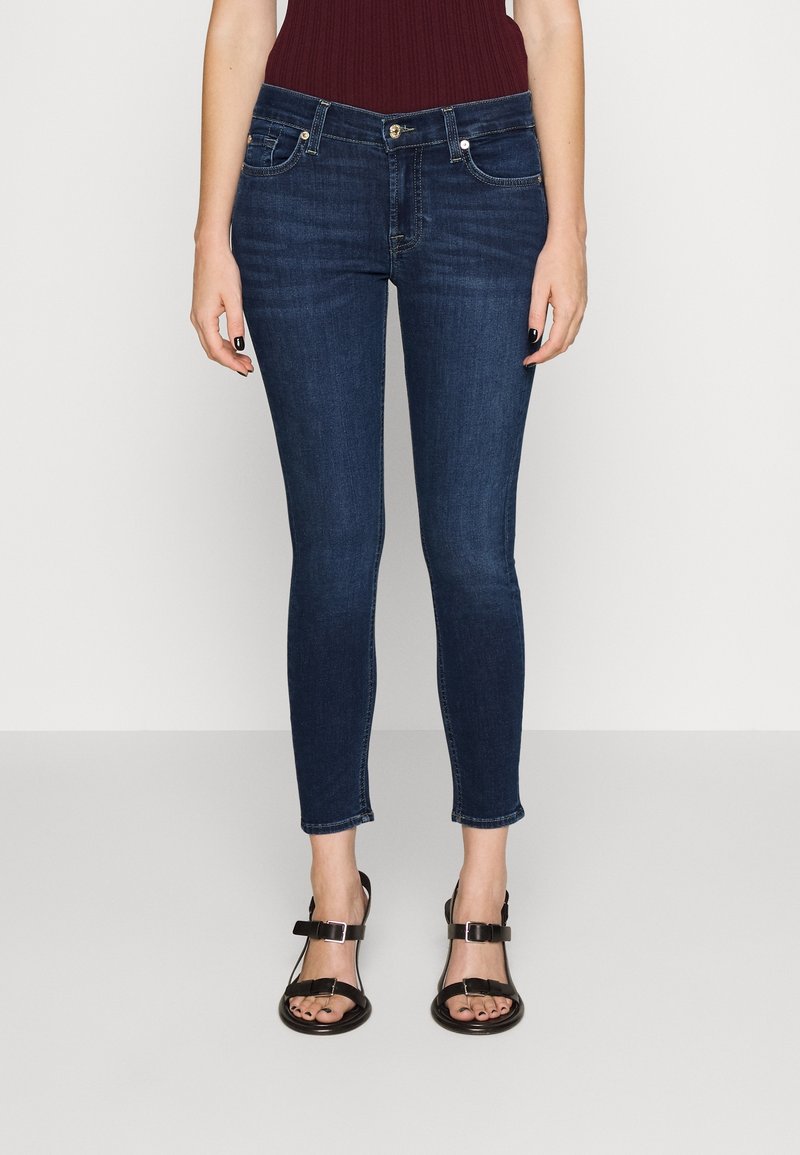 Dark blue skinny jeans made of denim, featuring front pockets, a zip fly, and a cropped length just above the ankle. Paired with black sandals.