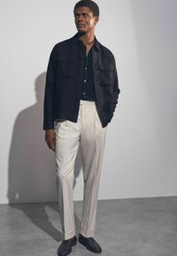Cămașă neagră cu nasturi, având două buzunare pe piept, asortată cu pantaloni crem croit și pantofi loafers întunecați, pe un fundal neutru.
