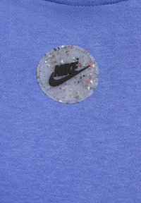 Violet medvilnės audinys turi apvalų, blizgantį etikėtę su juodu "NIKE" logotipu, dėmėtu dizainu ir siūlėmis.