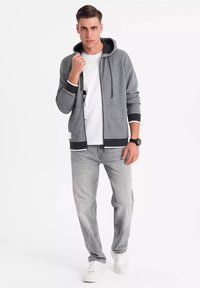 Sudadera gris con cremallera y acentos negros, llevada sobre una camiseta blanca, combinada con jeans gris claros y zapatillas blancas. Acabado de textil suave.