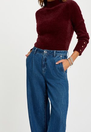 Femme portant un pull bordeaux en tissu duveteux à manches longues avec des boutons dorés sur les poignets et un jean bleu ample taille haute, les mains dans les poches.
