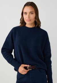 Maglione blu navy, morbido al tatto, con collo rotondo, polsini a coste e vestibilità ampia. Abbinato a pantaloni scuri e una cintura marrone con una fibbia dorata.
