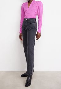 Henkilö seisoo pukeutuneena istuvaan vaaleanpunaiseen pitkähihaseen puseroon, korkeavyötäröisiin mustiin farkkuihin ja mustiin teräväkärkisiin nilkkureihin harmaalla lattialla.