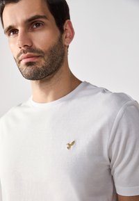 Fehér pamut póló, bal mellen egy apró, arany hímzett madárlogóval. Kerek nyakkivágás, rövid ujjú, sima tapintású.