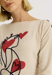 Pull beige avec un décolleté décontracté, orné de dessins en ligne noire et de cœurs rouges. Texture lisse avec des boutons noirs décoratifs sur les manches.