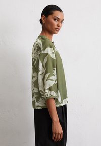 Green blouse with white floral pattern, round neckline, 3/4 sleeves, and gathered cuffs. Lightweight fabric, relaxed fit, paired with black pants.