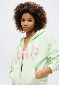 Νεαρή γυναίκα με σγουρά μαλλιά, φορώντας μια ανοιχτοπράσινη φούτερ GAP με φερμουάρ και λουλουδάτα γράμματα, με τα χέρια στις τσέπες, μπροστά σε απλό φόντο.