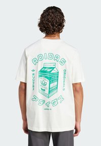 Fehér pamut póló, zöld grafikával, amelyen egy gyümölcslé carton látható japán felirattal: "ADIDAS ORIGINALS" és levél motívumok.