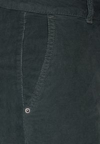 Gros plan sur la poche d'un pantalon en velours côtelé vert foncé avec des coutures visibles et un petit rivet métallique près du coin inférieur.