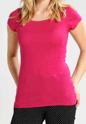 Frau, die ein eng anliegendes, leuchtend pinkes kurzärmliges Oberteil mit gezacktem Ausschnitt und schwarze Hosen mit weißen Punkten vor einem einfarbigen Hintergrund trägt.