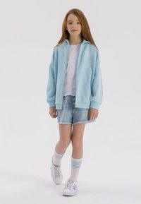 Svijetloplavi zip-up dukserica, bijela majica, oštećene traper šortsevi i bijele tenisice s plavim naglascima. Model nosi visokosnežne prugaste čarape.