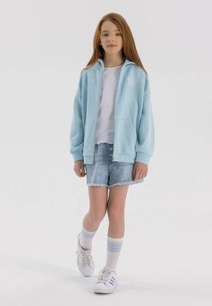 Felpa con zip azzurro chiaro, t-shirt bianca, pantaloni corti in denim strappati, e sneakers bianche con dettagli blu. Il modello indossa calze a righe sopra il ginocchio.