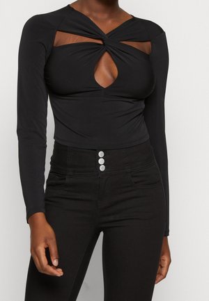 Sort langærmet bodysuit med vridset foran og nøglehuludskæring, parret med højtaljede sorte jeans med tre-knaps lukning og lommer.