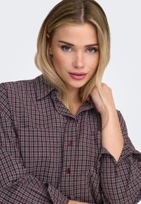 Chemise à carreaux rouge et gris avec col boutonné, manches longues, détail de poche et boutons rouges. Tissu doux avec une coupe décontractée.