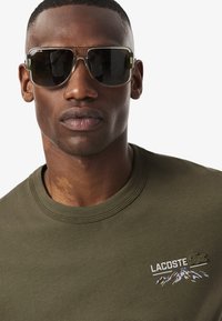 Camiseta de algodón verde con cuello redondo y logo bordado de Lacoste, además de un diseño de montaña. El modelo lleva unas grandes gafas de sol rectangulares con lentes oscuros.