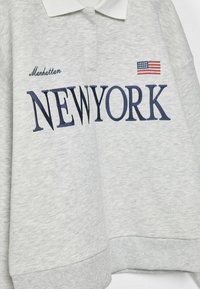 Sweat-shirt gris clair avec col blanc, texte brodé "Manhattan NEW YORK" et petit drapeau américain sur la poitrine.