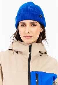 Beige hoodie met blauwe accenten en rits; ribgebreide blauwe muts. Glad oppervlak, functioneel ontwerp met zichtbare hardware.