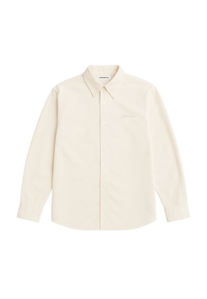 Uomo che indossa una camicia beige con bottoni e colletto e un berretto beige e nero su uno sfondo grigio uniforme.