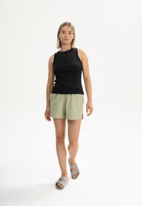 Schwarzes gestreiftes ärmelloses Oberteil, hellgrüne Shorts mit Kordelzug und seitlichen Schlitzen, graue Sandalen mit Schnallen. Schlichter Hintergrund.