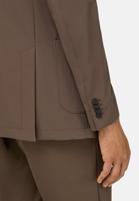 Ruskea räätälöity bleiseri, jossa on sileä pinta, sivutaskut ja neljä mustaa nappia hihassa. Muotoilu on tyylikäs, jossa on minimaalinen ompelu.