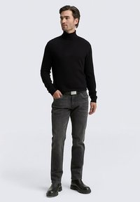 Schwarzer, enganliegender Rollkragenpullover; graue Denim-Jeans mit feiner Textur; schwarze Gummistiefel; legere Kleidung, stehende Pose, neutraler Hintergrund.