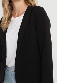 Černý saka na míru s vykrojeným límcem, nošené přes bílé tričko. Vyrobeno z hladké tkaniny a strukturovaného střihu, v kombinaci se světle modrými džíny.