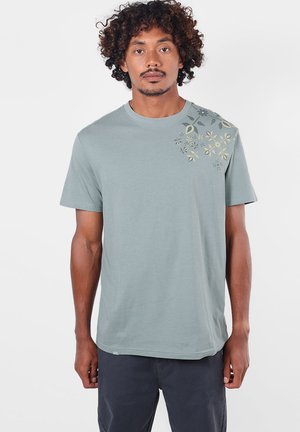 Homme aux cheveux bouclés portant un t-shirt gris-vert clair à manches courtes avec de petits motifs floraux sur l'épaule gauche.