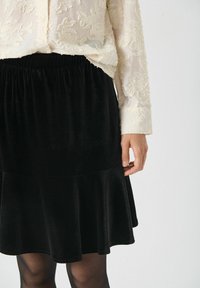 Zwarte fluwelen rok met een uitlopende ontwerp, elastische tailleband en zachte textuur. Gecombineerd met een crèmekleurige blouse met textuur.