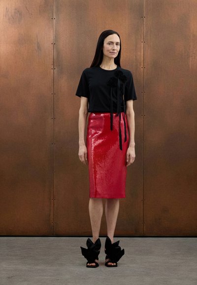 T-shirt noir avec accents floraux, assorti à une jupe rouge à sequins brillante mi-genoux et des sandales noires ornées de grands nœuds.