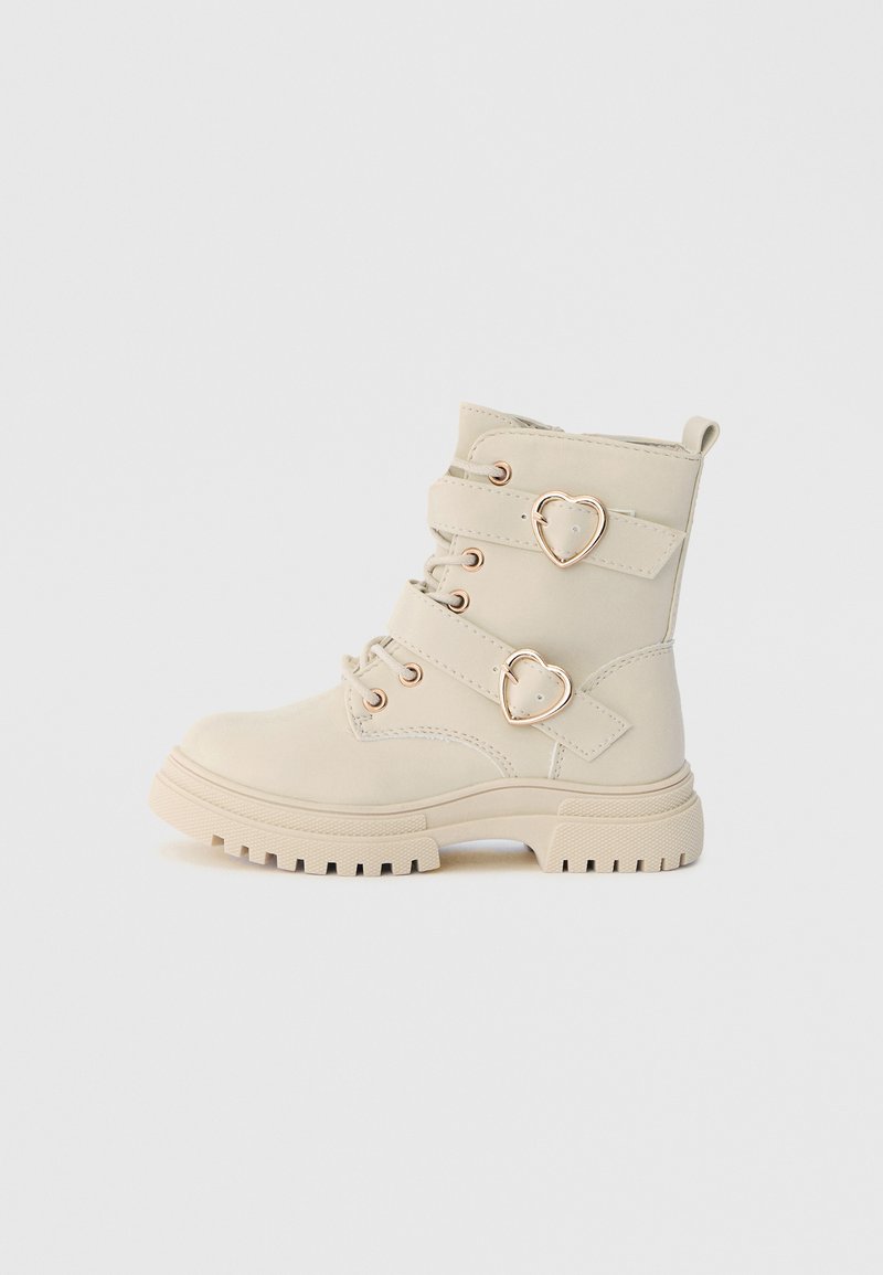 Bottes courtes beiges en matériau synthétique, avec double accent en forme de cœur, plusieurs sangles et une semelle en caoutchouc épaisse avec motif de bande de roulement.