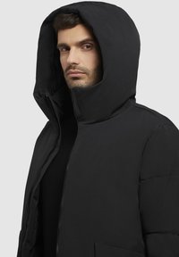 Chaqueta acolchada negra con una gran capucha, cierre delantero y bolsillos laterales. La tela parece suave y aislante, diseñada para brindar calor.