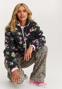 Fleece bunda s tmavým pozadím a kvetinovými vzormi v ružovej a zelenej farbe, skombinovaná s nohavicami s leopardím vzorom a ružovými topánkami so pruhmi.