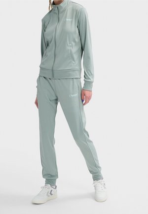 Osoba ubrana w jasno szaro-zieloną bluzę sportową na zamek i pasujące spodnie oraz białe tenisówki, stojąca na tle jednolitego tła.