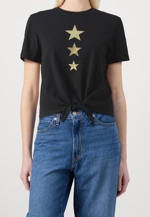 Black short-sleeve shirt with three descending gold stars on front, knotted at waist, worn with high-waisted blue jeans.