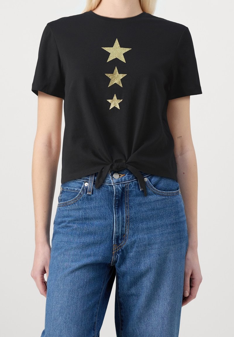 Chemise noire à manches courtes avec trois étoiles dorées décroissantes sur le devant, nouée à la taille, portée avec un jean bleu taille haute.