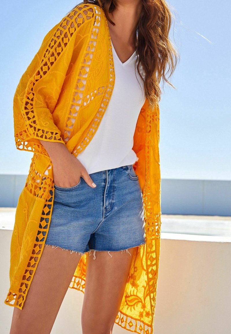 Femme portant un t-shirt blanc col en V, un short en jean bleu et un long cardigan jaune en dentelle, debout dehors avec une main dans la poche.