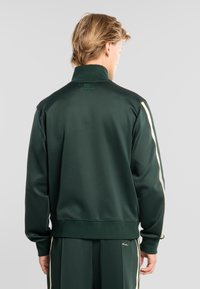 Hombre con chándal verde oscuro con franjas claras en las mangas y los pantalones, de pie de espaldas contra un fondo liso.