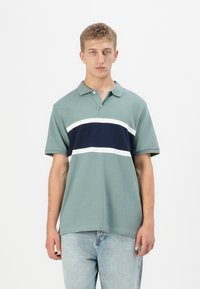 Mladenič oblečen v potisnjeno zeleno polo majico s temno modrimi in belimi vodoravnimi črtami, v kombinaciji z svetlo modrimi jeans hlačami, stoji na beli podlagi.