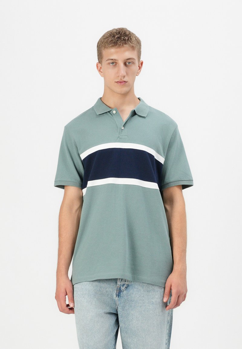 Mladenič oblečen v potisnjeno zeleno polo majico s temno modrimi in belimi vodoravnimi črtami, v kombinaciji z svetlo modrimi jeans hlačami, stoji na beli podlagi.