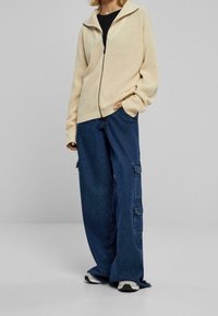 Světlý béžový svetr na zip s žebrovanou texturou a spadlými rameny, kombinovaný s volně střiženými tmavě modrými cargo džínami s bočními kapsami.