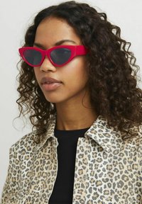 Red, cat-eye sunglasses with a solid frame and dark lenses, worn over a leopard-print shirt, highlighting bold design and contrasting colors.