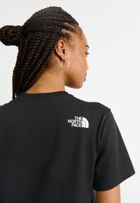 Tricou negru din bumbac, cu mâneci scurte, având un logo alb pe partea superioară a spatelui. Textură netedă și croială lejeră.