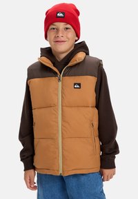 Pojke med röd basker, brun hoodie och en tan- och brun pufferväst med dragkedja fram och logopatch, står mot vit bakgrund.