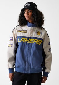 Blue and cream jacket featuring "LAKERS" text, various patches, and contrasting black sleeves. Ribbed cuffs and collar details.
