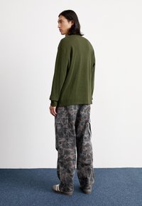 Sweter w kolorze oliwkowym z fakturą, szare spodnie cargo w kamuflażu z wieloma kieszeniami i ciemne sandały. Model widoczny od tyłu. Tło białej ściany.