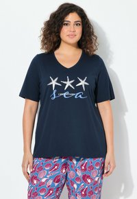 T-shirt navy a scollo a V con grafica di stella marina argento e testo "Ho bisogno di vitamina mare". Abbinato a pantaloni con fantasia di conchiglie su sfondo blu.